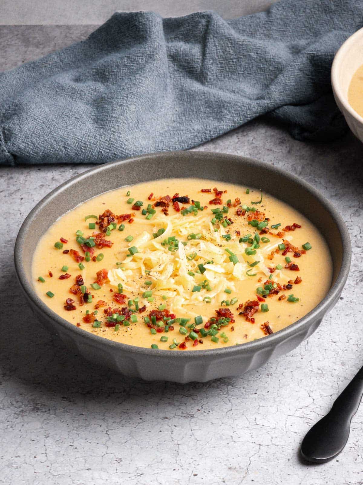A bowl of potato leek soup (with bacon) on a concrete counter top topped with bacon, shredded gouda, and chives.