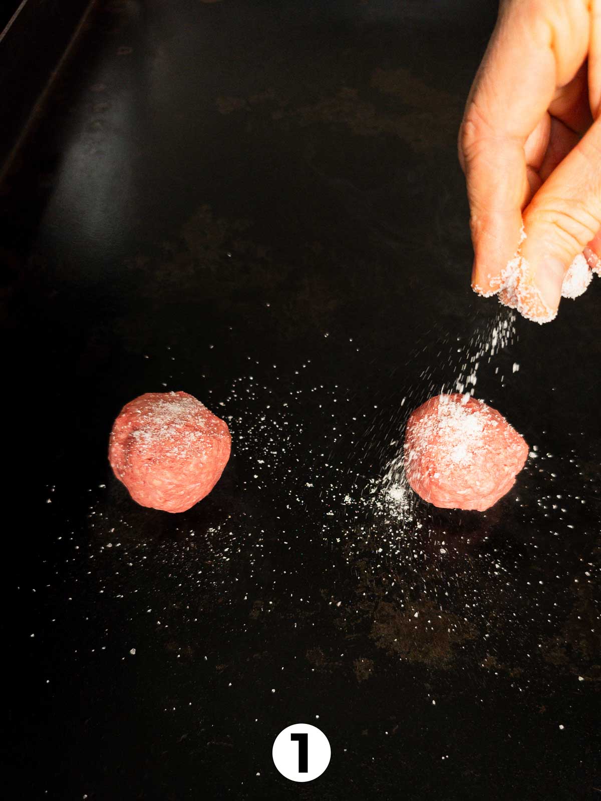 Two balls of 80/20 ground beef being salted on a hot griddle for smash burgers.