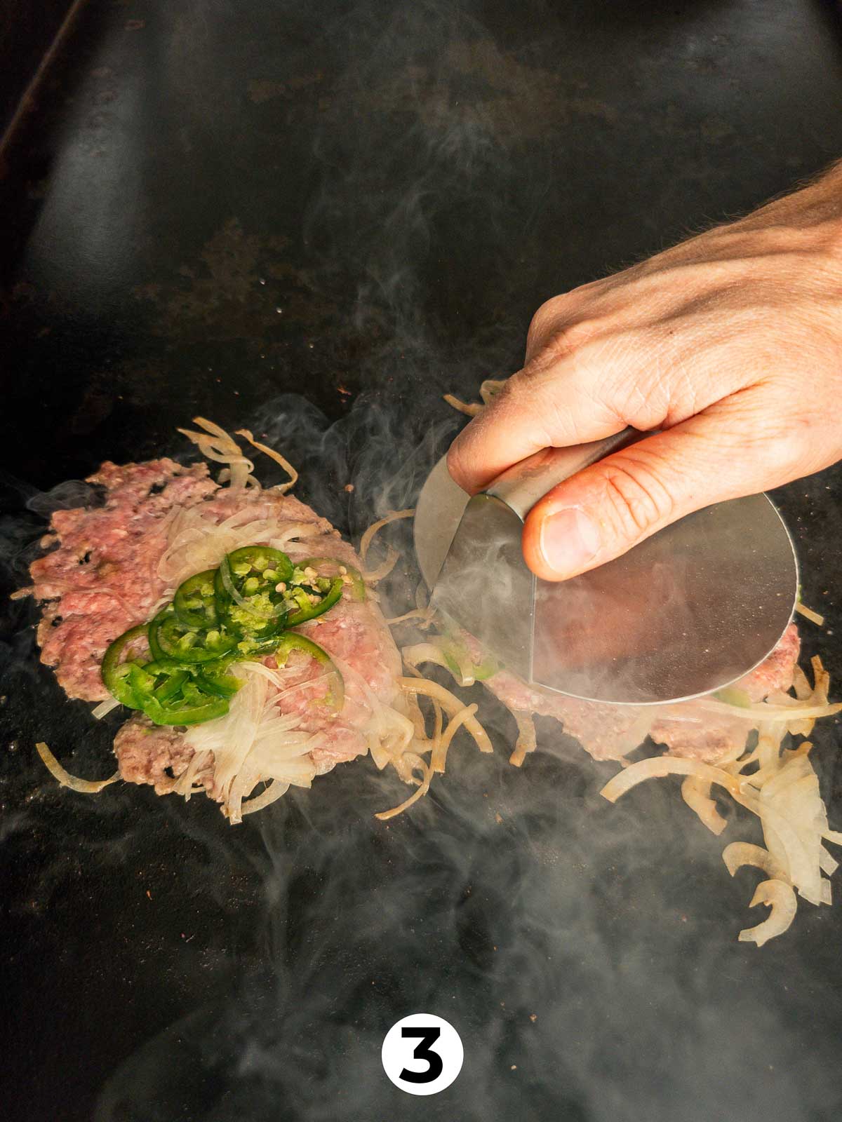 A hand using a burger press to smash balls of ground beef covered in onions and jalapeño slices into burger patties.